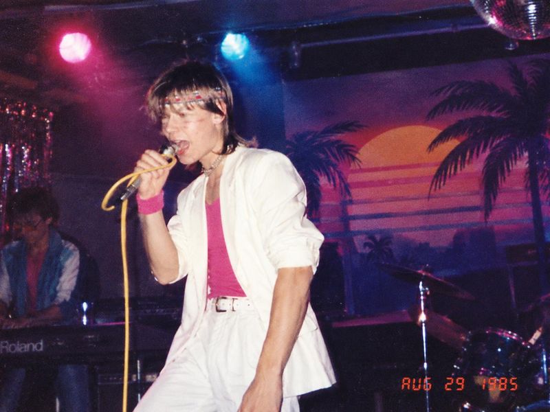 Tommy Cape singing on stage in a white suit, pink top and headband, a keyboard player and a palm-tree sunset backdrop behind him.